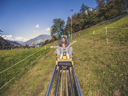 Dein Apartment im Zillertal in bester Lage Mann und Kind fahren Rodelbahn auf Gras mit Bergen im Hintergrund